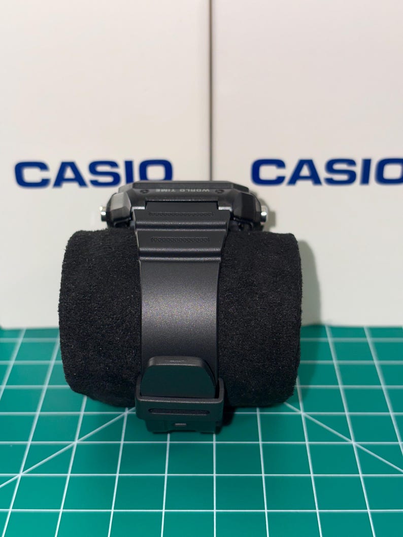 Peut inclure: Une montre num&eacute;rique Casio noire avec un bracelet noir est pr&eacute;sent&eacute;e sur un coussin cylindrique noir. La montre a un cadran carr&eacute; et une fermeture &agrave; boucle. Le mot "CASIO" est visible en arri&egrave;re-plan.