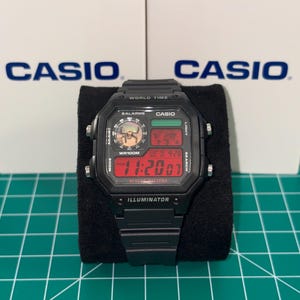 Puede incluir: Reloj digital Casio negro con una esfera rectangular que muestra la hora en d&iacute;gitos rojos. El reloj tiene m&uacute;ltiples botones y funciones, incluyendo "WORLD TIME" e "ILLUMINATOR". El logotipo de Casio es visible en la esfera y en el fondo.