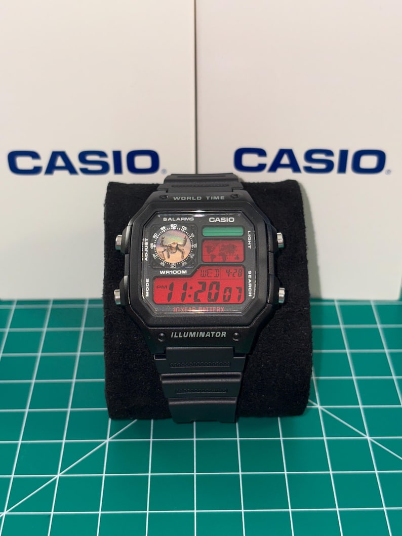 Puede incluir: Reloj digital Casio negro con una esfera rectangular que muestra la hora en d&iacute;gitos rojos. El reloj tiene m&uacute;ltiples botones y funciones, incluyendo "WORLD TIME" e "ILLUMINATOR". El logotipo de Casio es visible en la esfera y en el fondo.