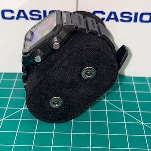 May include: Black digital watch with a rectangular face and a black band. The watch is displayed on a black velvet stand. The background features the word "CASIO" in blue letters.