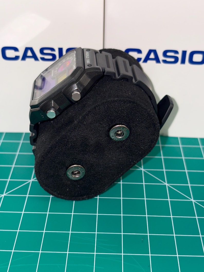 May include: Black digital watch with a rectangular face and a black band. The watch is displayed on a black velvet stand. The background features the word "CASIO" in blue letters.