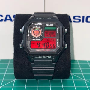 May include: Black digital watch with a rectangular face. The watch displays the time in red digits and features the text "WORLD TIME", "SALARMS", "CASIO", and "ILLUMINATOR". The watch has a black band and buttons.