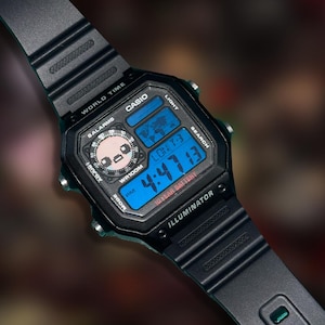 Può includere: Orologio digitale Casio nero con display digitale blu che mostra l'ora e un'icona della mappa del mondo. L'orologio ha un cinturino nero e presenta il testo "CASIO", "WORLD TIME" e "ILLUMINATOR".
