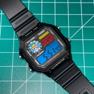 K&ouml;nnte beinhalten: Schwarze digitale Casio-Uhr mit rechteckigem Zifferblatt. Die Uhr zeigt die Uhrzeit, das Datum und andere Funktionen auf einem blauen Digitalbildschirm an. Das Zifferblatt der Uhr tr&auml;gt die Aufschrift "CASIO", "WR100M" und "ILLUMINATOR". Das Armband der Uhr ist schwarz.