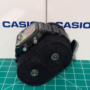 Peut inclure: Montre num&eacute;rique Casio noire avec un cadran rectangulaire et un bracelet noir. La montre est pr&eacute;sent&eacute;e sur un support en velours noir. Le mot "CASIO" est visible en arri&egrave;re-plan.
