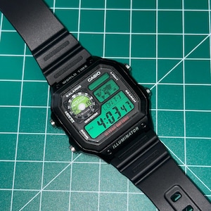 May include: Black Casio digital watch with a rectangular face and a black band. The watch face displays the time in green digits, along with the day and date. The watch has multiple function buttons and the text "ILLUMINATOR".