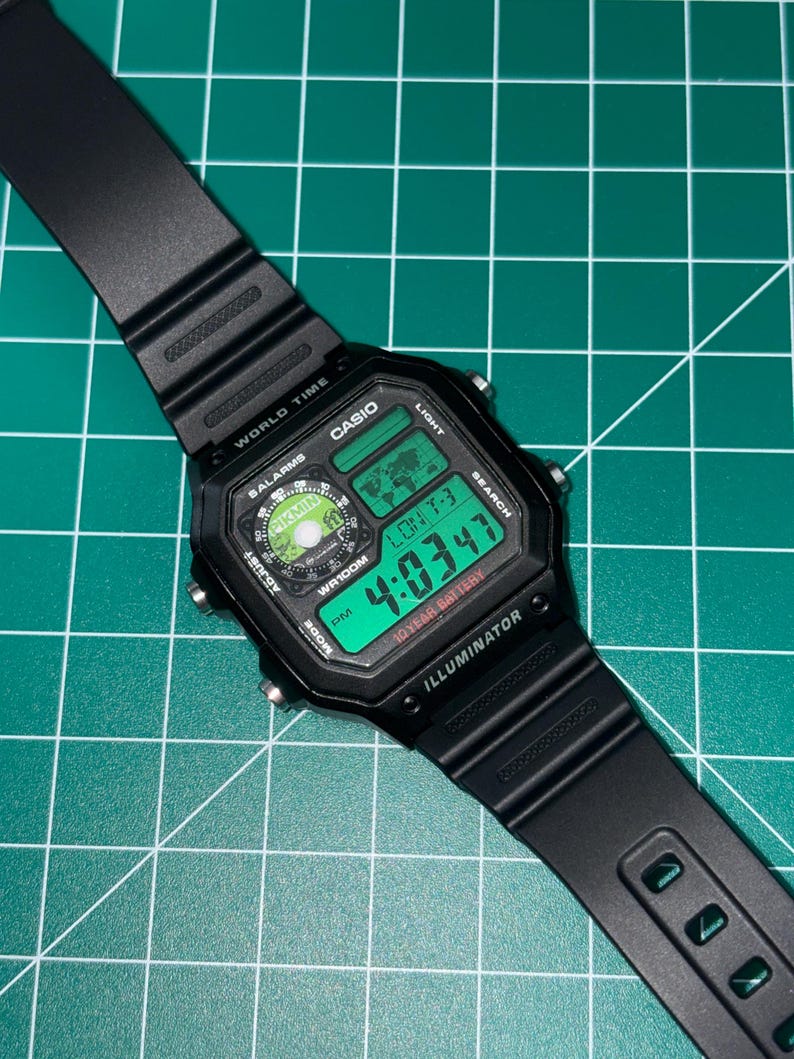 May include: Black Casio digital watch with a rectangular face and a black band. The watch face displays the time in green digits, along with the day and date. The watch has multiple function buttons and the text "ILLUMINATOR".