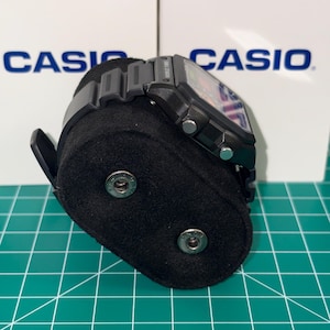 Puede incluir: Un reloj digital negro con una esfera rectangular se muestra en un soporte de reloj negro. El reloj tiene una correa negra y botones plateados. El fondo presenta la palabra "CASIO" en blanco.