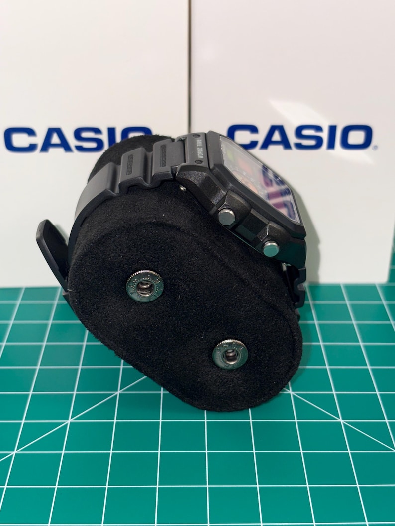Puede incluir: Un reloj digital negro con una esfera rectangular se muestra en un soporte de reloj negro. El reloj tiene una correa negra y botones plateados. El fondo presenta la palabra "CASIO" en blanco.
