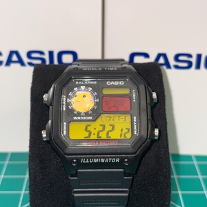 May include: Black Casio digital watch with a rectangular face. The watch displays the time, date, and world time functions. The face has a yellow digital display with the text "ILLUMINATOR" at the bottom. The watch is on a black surface.