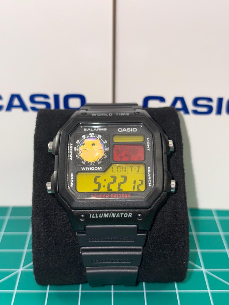 May include: Black Casio digital watch with a rectangular face. The watch displays the time, date, and world time functions. The face has a yellow digital display with the text "ILLUMINATOR" at the bottom. The watch is on a black surface.