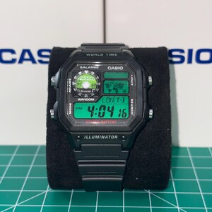 May include: Black digital watch with a rectangular face, displaying the time 4:04:16 PM. The watch features a green digital display, with the word "ILLUMINATOR" at the bottom. The watch is on a black display stand.