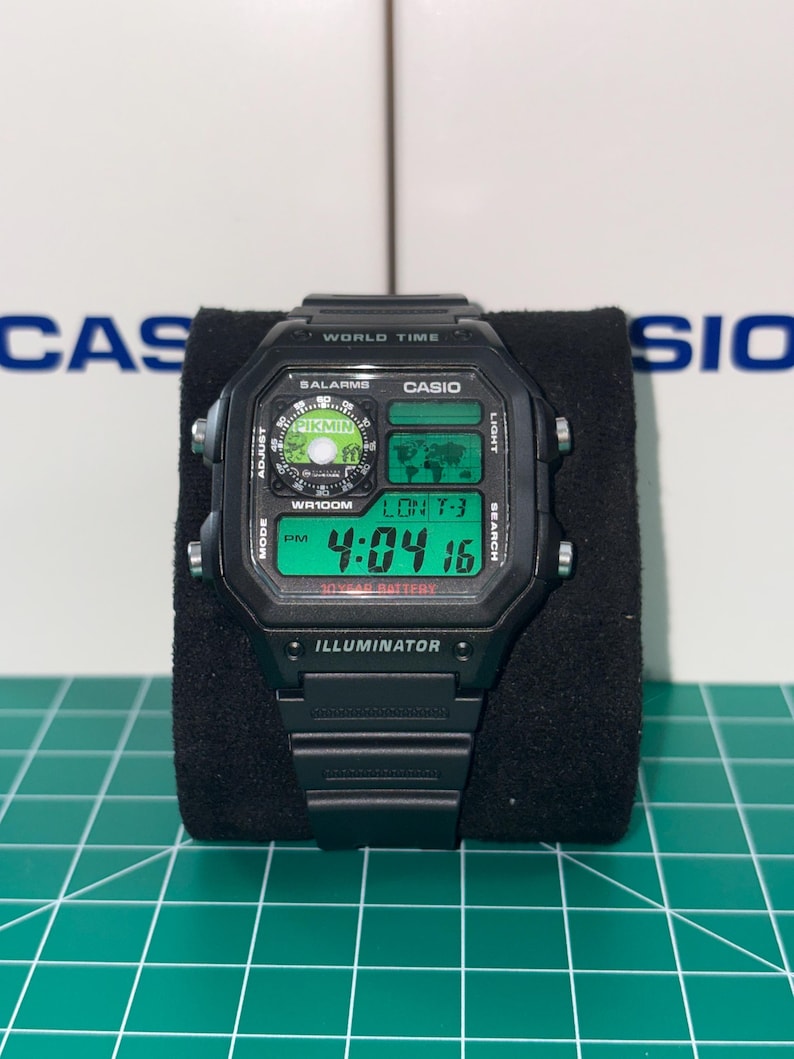 May include: Black digital watch with a rectangular face, displaying the time 4:04:16 PM. The watch features a green digital display, with the word "ILLUMINATOR" at the bottom. The watch is on a black display stand.