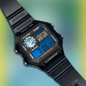 Può includere: Orologio digitale Casio nero con display digitale blu che mostra l'ora. L'orologio presenta una grafica della mappa del mondo e il testo "WORLD TIME", "CASIO" e "ILLUMINATOR". Il cinturino dell'orologio è nero.