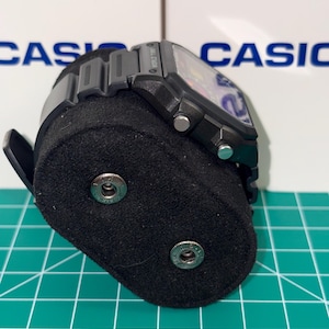 May include: A black digital watch with a rectangular face and a black band. The watch is displayed on a black velvet watch stand. The word "CASIO" is visible in the background.