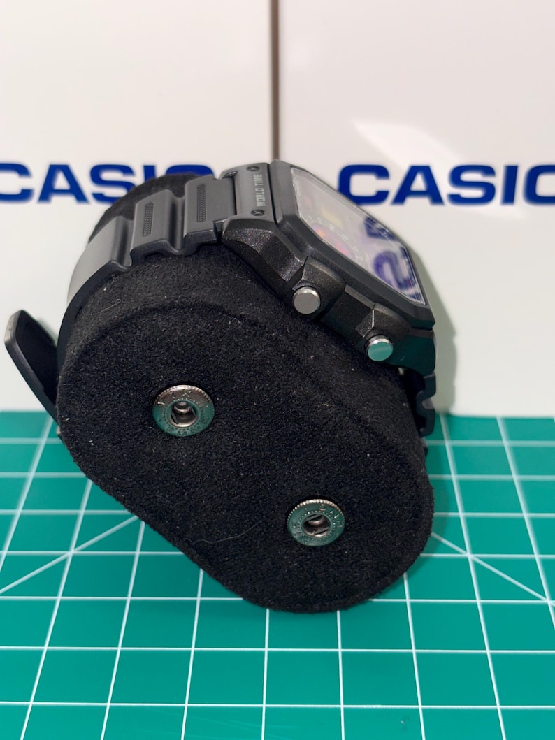 May include: A black digital watch with a rectangular face and a black band. The watch is displayed on a black velvet watch stand. The word "CASIO" is visible in the background.