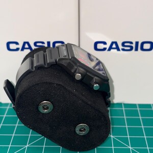 Peut inclure: Une montre num&eacute;rique Casio noire avec un cadran rectangulaire et un bracelet noir. La montre est pr&eacute;sent&eacute;e sur un support de montre noir. Le mot "CASIO" est visible en arri&egrave;re-plan.