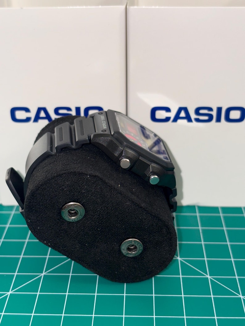 Peut inclure: Une montre num&eacute;rique Casio noire avec un cadran rectangulaire et un bracelet noir. La montre est pr&eacute;sent&eacute;e sur un support de montre noir. Le mot "CASIO" est visible en arri&egrave;re-plan.