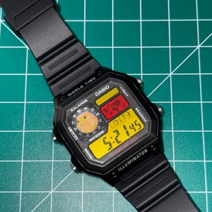 May include: Black Casio digital watch with a square face and black band. The watch face displays the time in yellow digits, along with other functions. The watch has the text "CASIO", "WORLD TIME", and "ILLUMINATOR".