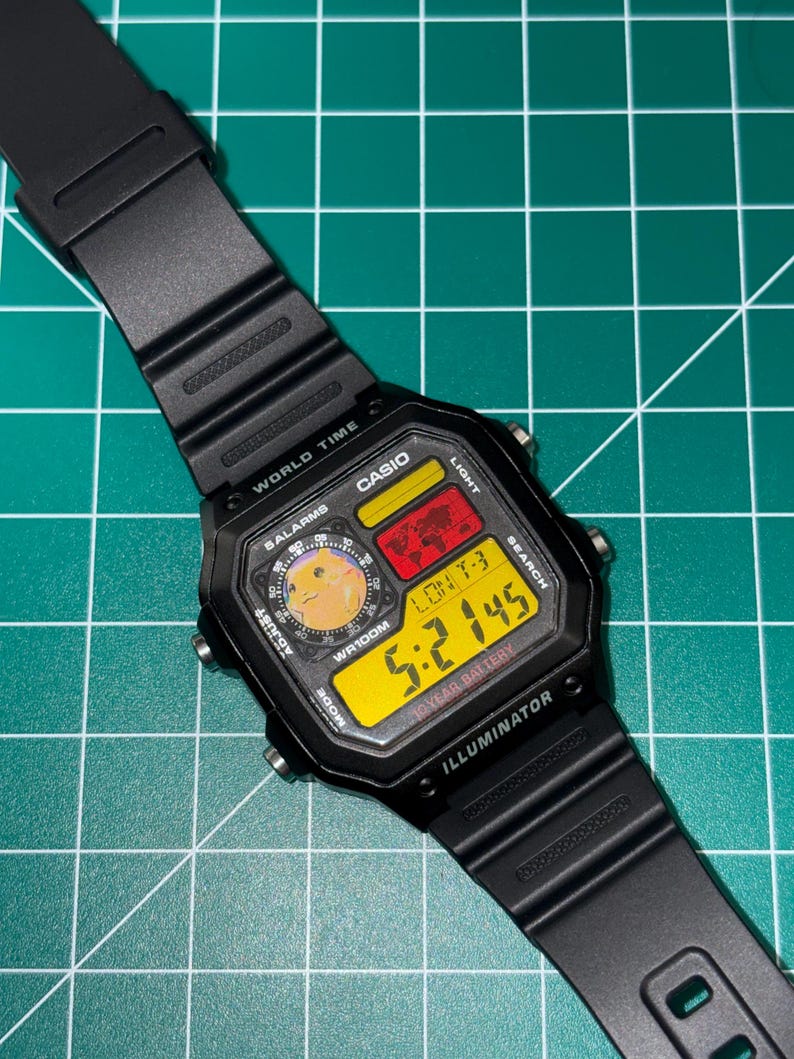May include: Black Casio digital watch with a square face and black band. The watch face displays the time in yellow digits, along with other functions. The watch has the text "CASIO", "WORLD TIME", and "ILLUMINATOR".