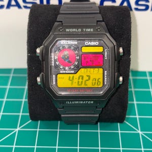 Peut inclure: Montre num&eacute;rique Casio noire avec un cadran rectangulaire. La montre affiche l'heure, la date et les fonctions de l'heure mondiale. Le cadran a des accents jaunes, rouges et noirs, avec le texte "WORLD TIME" et "ILLUMINATOR".