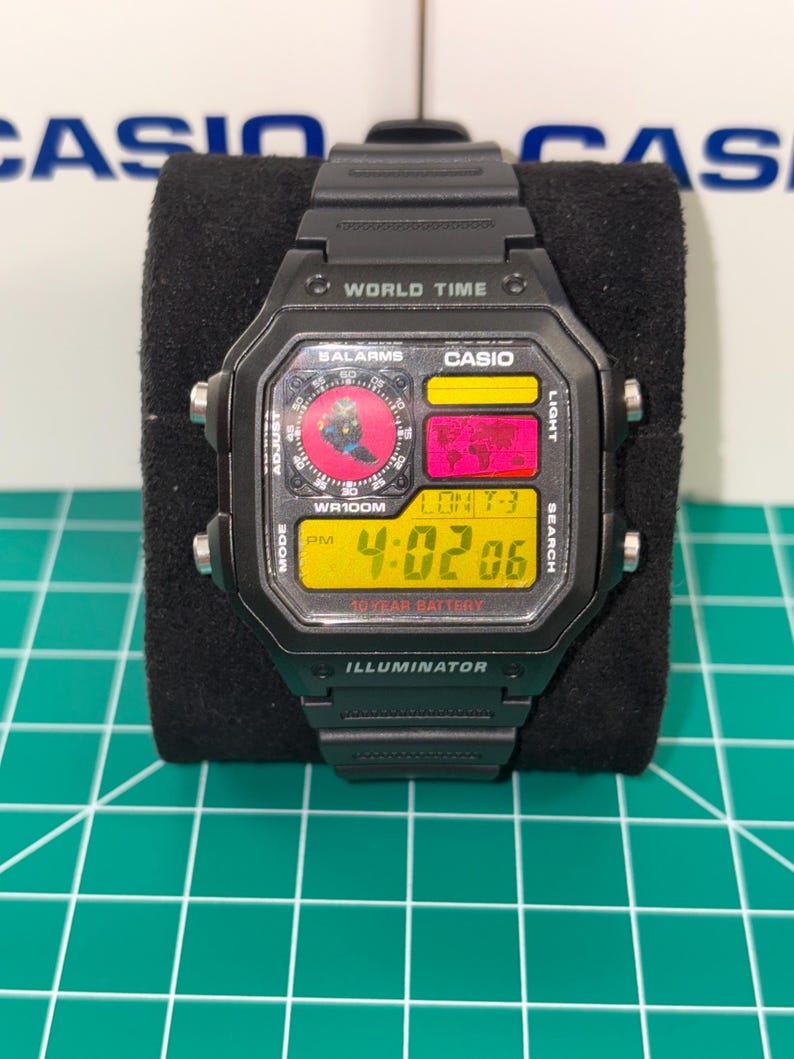 Peut inclure: Montre num&eacute;rique Casio noire avec un cadran rectangulaire. La montre affiche l'heure, la date et les fonctions de l'heure mondiale. Le cadran a des accents jaunes, rouges et noirs, avec le texte "WORLD TIME" et "ILLUMINATOR".