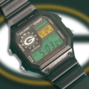 Könnte beinhalten: Eine schwarze Casio Digitaluhr mit geripptem Kunststoffarmband. Das Zifferblatt zeigt eine Weltkarte, ein kreisförmiges Feld mit grünem G-Logo und ein Digitaldisplay mit TUE 4-20 und 5:03:04. Aufschrift: WORLD TIME, 5 ALARMS, WR100M, ILLUMINATOR.