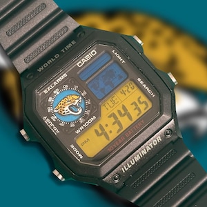 Könnte beinhalten: Eine schwarze digitale Casio-Armbanduhr mit Kunstharzarmband, rechteckigem Gehäuse, gelbem Display, Weltkarte und dem Logo der Jacksonville Jaguars auf dem runden Zifferblatt. Das Display zeigt die Uhrzeit 4:34:35 und das Datum TUE 4-20.