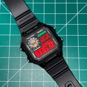 Puede incluir: Reloj digital Casio negro con esfera rectangular y correa negra. La esfera del reloj muestra la hora en d&iacute;gitos rojos, junto con el d&iacute;a y la fecha. El reloj tiene m&uacute;ltiples botones y funciones, incluyendo hora mundial y alarmas.