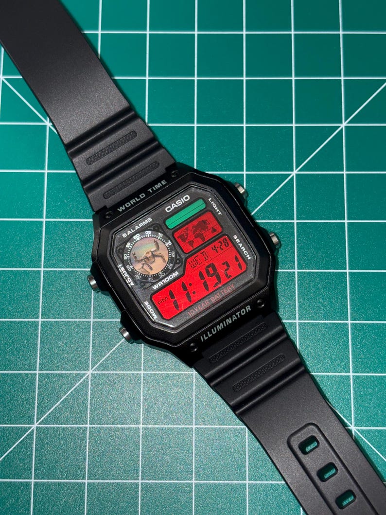 Puede incluir: Reloj digital Casio negro con esfera rectangular y correa negra. La esfera del reloj muestra la hora en d&iacute;gitos rojos, junto con el d&iacute;a y la fecha. El reloj tiene m&uacute;ltiples botones y funciones, incluyendo hora mundial y alarmas.