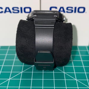 Puede incluir: Un reloj digital Casio negro con una esfera cuadrada y una correa negra. El reloj se muestra sobre un coj&iacute;n cil&iacute;ndrico negro. La palabra "CASIO" es visible en el fondo.