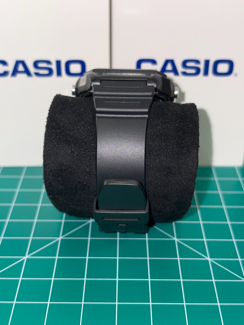 Puede incluir: Un reloj digital Casio negro con una esfera cuadrada y una correa negra. El reloj se muestra sobre un coj&iacute;n cil&iacute;ndrico negro. La palabra "CASIO" es visible en el fondo.
