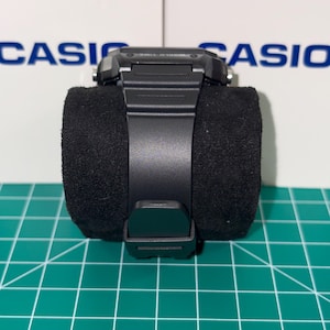 May include: A black digital wristwatch with a black band is displayed on a black velvet cushion. The watch has a rectangular face and a buckle closure. The background features the word "CASIO" in blue.
