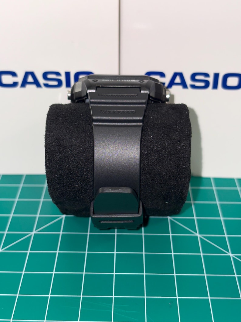 May include: A black digital wristwatch with a black band is displayed on a black velvet cushion. The watch has a rectangular face and a buckle closure. The background features the word "CASIO" in blue.