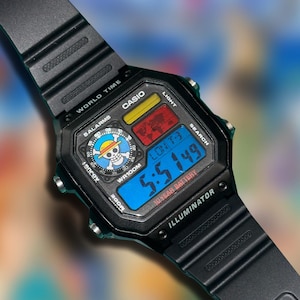 K&ouml;nnte beinhalten: Schwarze digitale Casio-Uhr mit blauem Digitaldisplay, das die Uhrzeit anzeigt. Das Zifferblatt zeigt ein Totenkopf- und gekreuzte Knochen-Logo mit Strohhut. Das Armband ist schwarz.