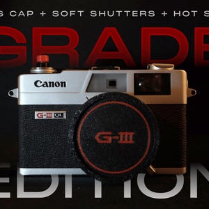 Puede incluir: Una cámara Canon G-III QL vintage con un cuerpo negro y plateado y una tapa de lente. La tapa de la lente tiene el texto "G-III" en rojo. La palabra "GRADE" está en letras rojas grandes encima de la cámara, y "EDITION" debajo.