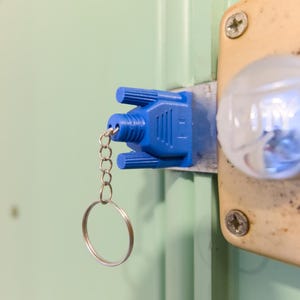 May include: A blue key cover attached to a key ring, inserted into a door lock. The lock is on a light-colored door, with a metal plate and a round, clear knob. The key cover has a unique, geometric design.