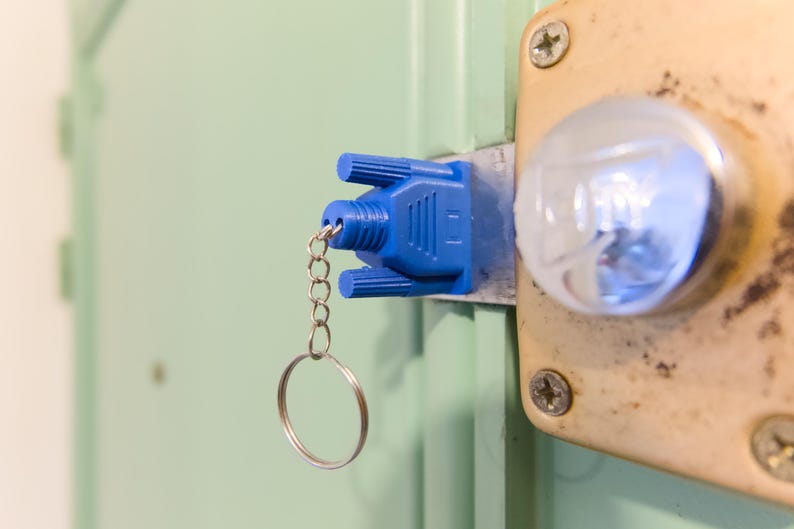May include: A blue key cover attached to a key ring, inserted into a door lock. The lock is on a light-colored door, with a metal plate and a round, clear knob. The key cover has a unique, geometric design.