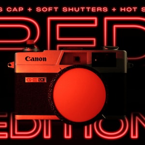 Puede incluir: Una cámara Canon G-III QL vintage con una tapa de lente roja, sobre un fondo negro. Las palabras "RED EDITION" están iluminadas en neón rojo. La cámara es negra y plateada.