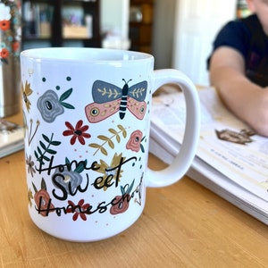 May include: White ceramic mug with a wraparound floral and butterfly design. The mug features a butterfly with pink and blue wings, along with flowers in shades of red, gray, and gold. The words "Home Sweet Homeschool" are written on the mug.