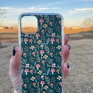 May include: Clear phone case with a dark green background featuring a pattern of red and white mushrooms, pink and yellow flowers, and green leaves. The case is held up against a blurred outdoor background.