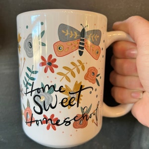 May include: White ceramic mug with a wraparound floral design and a large butterfly illustration. The mug features the words "Home Sweet Homeschool" in black script. The mug is held by a hand.