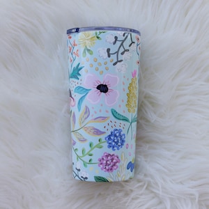 May include: A floral-patterned tumbler with a light blue background. The design features pink, purple, yellow, and blue flowers with green leaves. The tumbler has a clear lid and is likely made of stainless steel.