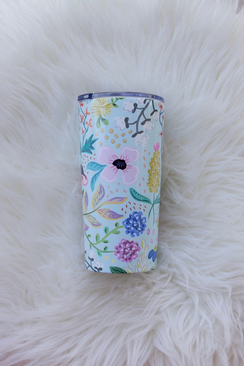May include: A floral-patterned tumbler with a light blue background. The design features pink, purple, yellow, and blue flowers with green leaves. The tumbler has a clear lid and is likely made of stainless steel.