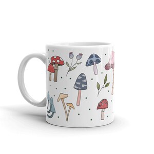 May include: White ceramic mug featuring a whimsical mushroom design. The design includes various colorful mushroom illustrations, including red, blue, and tan mushrooms, along with green polka dots and floral accents. The mug has a white handle.