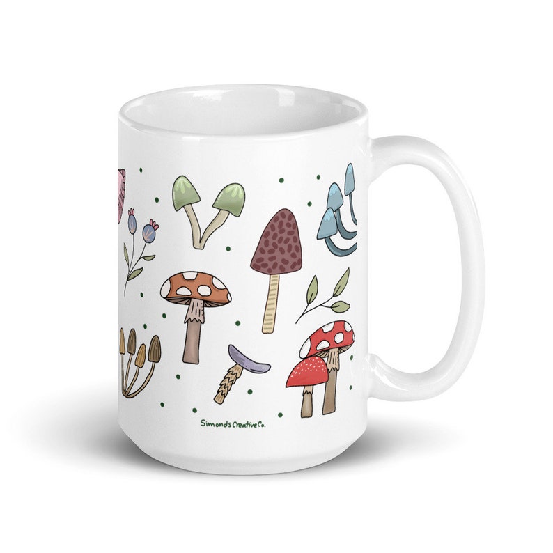 May include: White ceramic mug featuring a whimsical mushroom design in various colors, including red, brown, and green. The mug has a curved handle and the text "Simond's Creative Co." is visible on the side. The mug is approximately 4.5 inches tall.