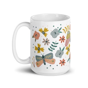 May include: White ceramic mug decorated with colorful butterflies and flowers in shades of pink, yellow, blue, and brown. The mug has a curved handle and a smooth, glossy finish. The design is whimsical and nature-themed.