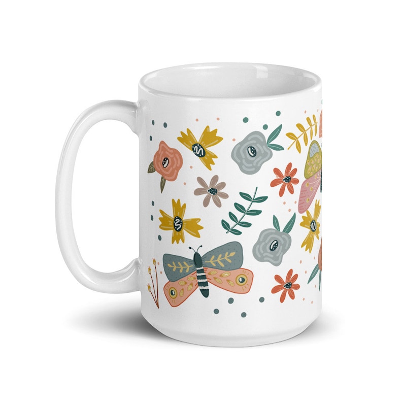 May include: White ceramic mug decorated with colorful butterflies and flowers in shades of pink, yellow, blue, and brown. The mug has a curved handle and a smooth, glossy finish. The design is whimsical and nature-themed.