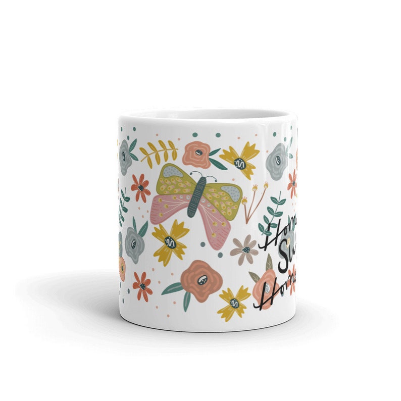 May include: White ceramic mug decorated with a colorful floral and butterfly design. The mug features a large butterfly with pink and yellow wings, surrounded by flowers in various colors. The words "Home Sweet Home" are written on the side.