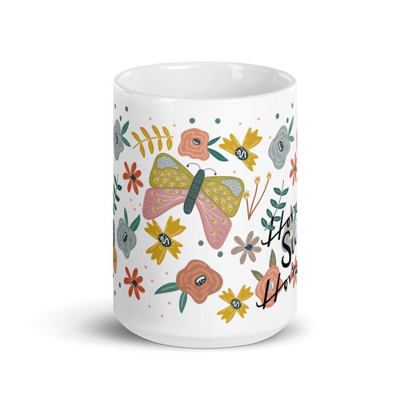 May include: White ceramic mug decorated with a colorful floral and butterfly pattern. The design includes pink, yellow, and orange flowers, a butterfly, and the words "Home Sweet Home" in black script. A cheerful, decorative item.